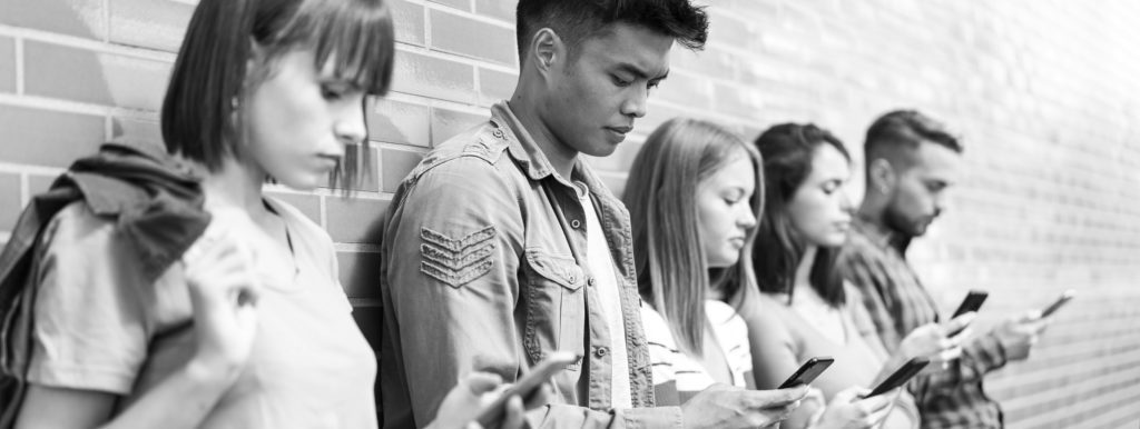 Five young people leaning against a wall all staring at phones looking neutral