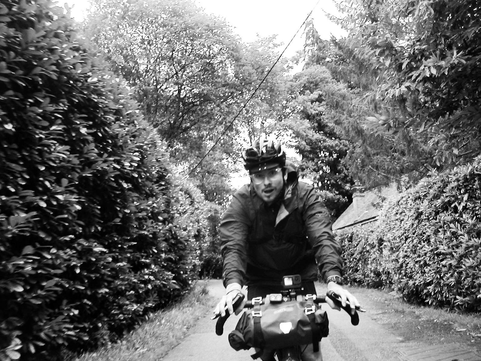 Black and white image of man cycling along narrow road surrounded by hedges and trees.