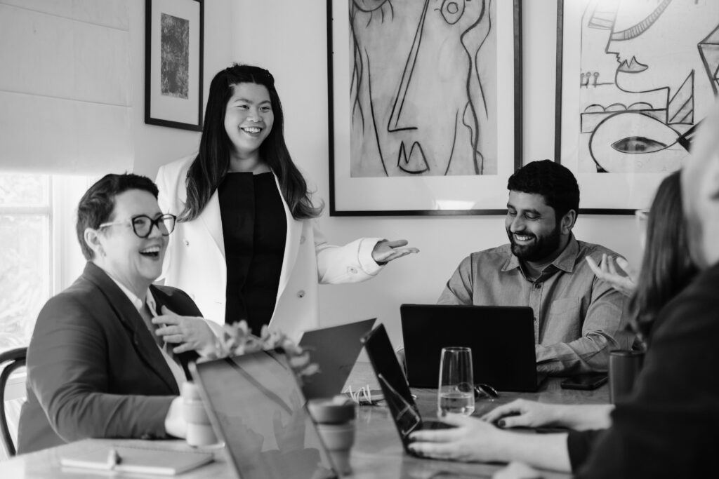 A group of colleagues smile as they meet around a table with their laptops. A woman and a man are seated and another woman is standing and gesturing to the group.