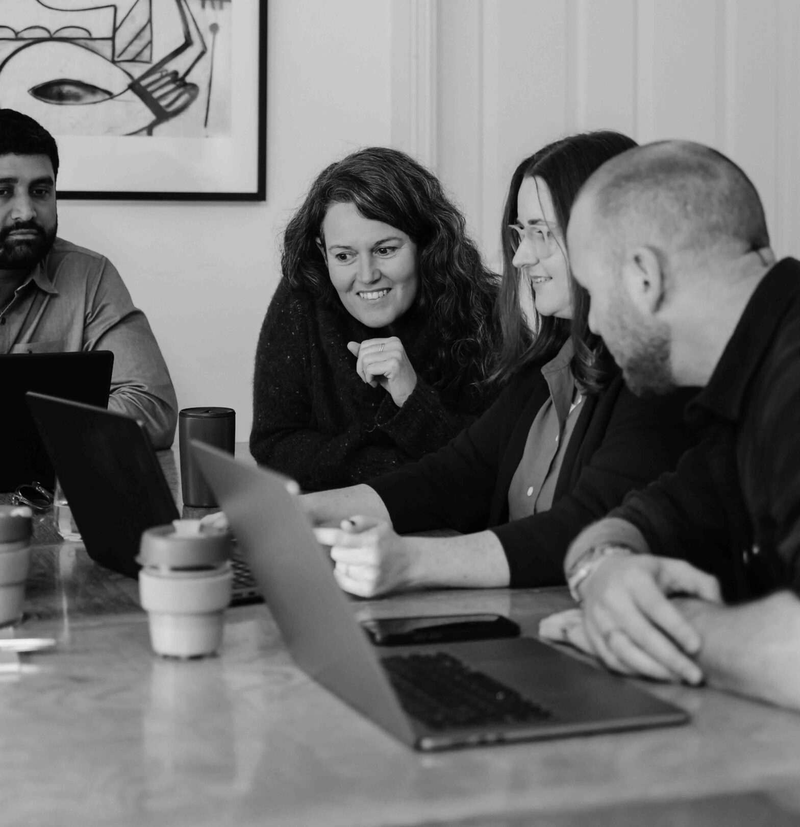 A group of four colleagues meet around a table with their laptops. All are seated and looking towards the same screen.